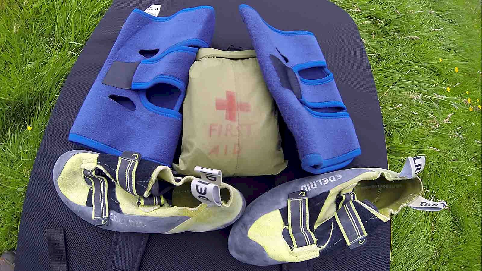 Picture of two knee support bandages with a pair of aggressive climbing shoes and a personal first aid kit spread out on a bouldering pad.